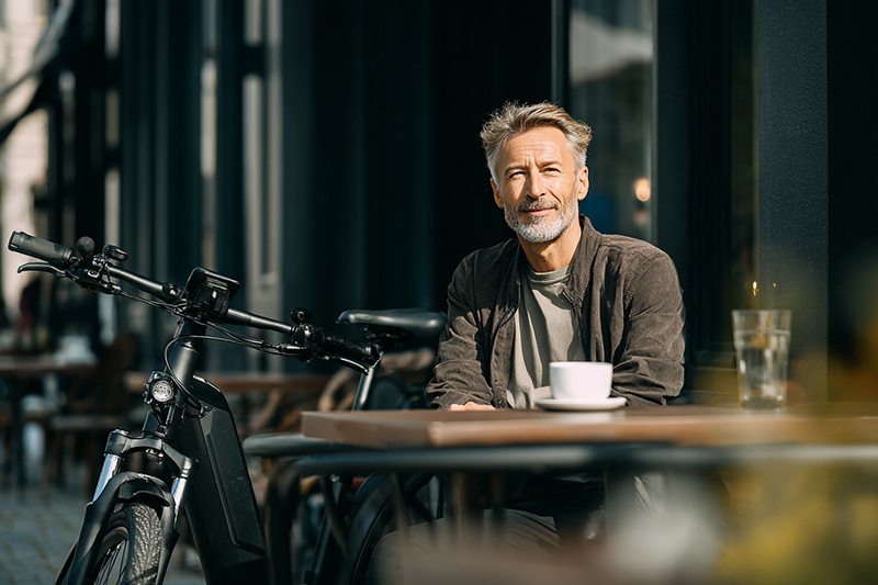 Zufriedener Mann genießt eine Kaffeepause in der Stadt, sein E-Bike ist dank einer Versicherung sicher neben ihm abgestellt.